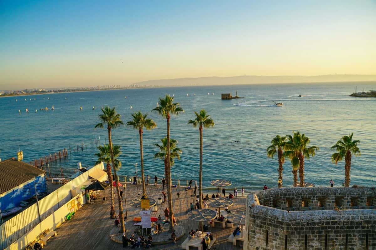 טיילת עם עצי דקל ליד חומת אבן עתיקה, ומולה מפרץ רחב והרים באופק בשעת שקיעה